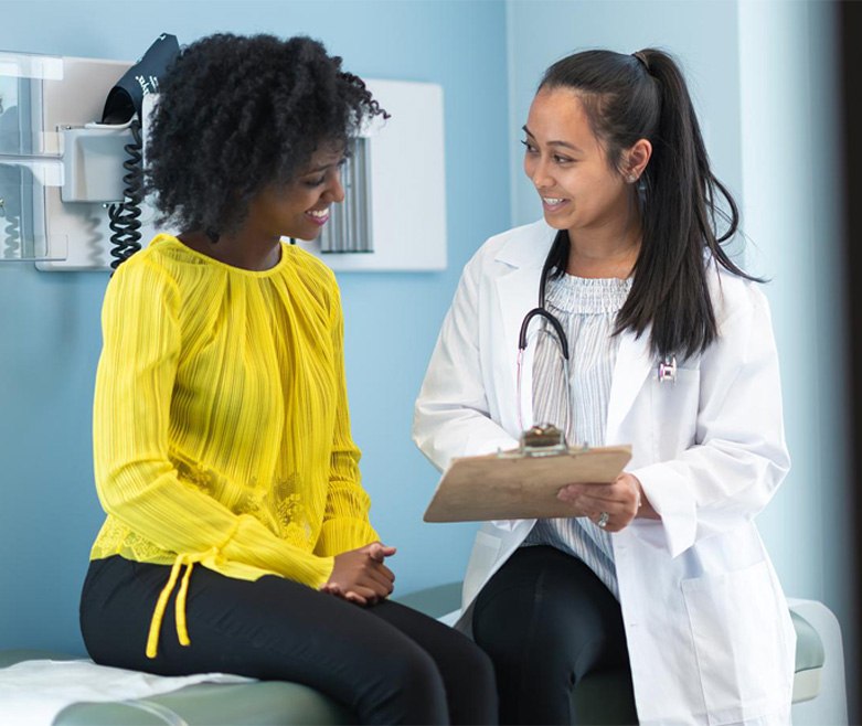 A young black woman is at a routine medical appointment. Her healthcare provider is an Asian woman. The patient and doctor are sitting next to each other on an examination table in a clinic. The doctor is asking the patient questions and taking notes on a clipboard.
